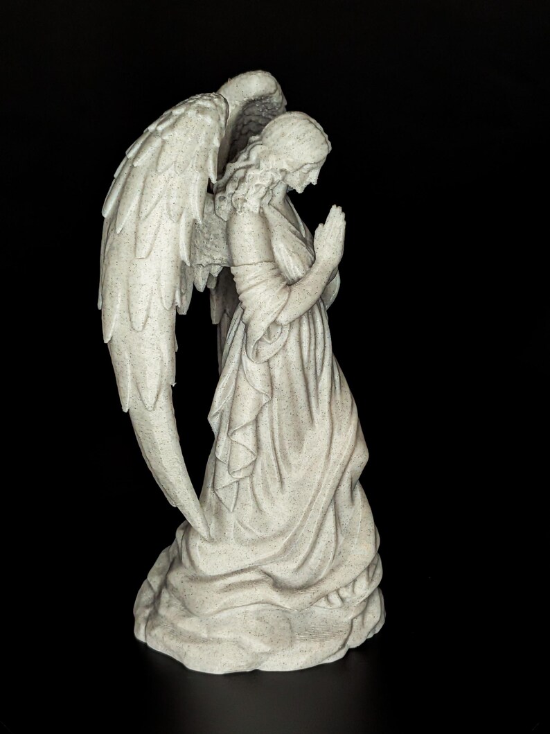 May include: A stone-colored angel statue with large, detailed wings. The angel is depicted in a praying pose, wearing a flowing gown. The statue has a textured surface and is set against a black background.