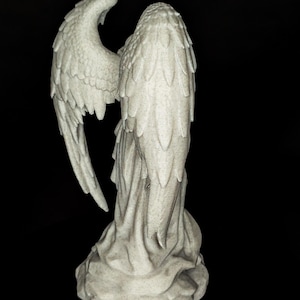 May include: A detailed, gray angel statue with outstretched wings. The sculpture features intricate feather detailing on the wings and a flowing robe. The statue is set against a black background, highlighting its form.