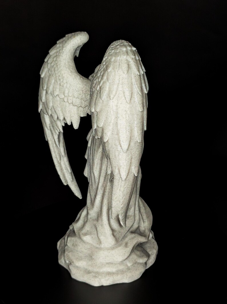 May include: A detailed, gray angel statue with outstretched wings. The sculpture features intricate feather detailing on the wings and a flowing robe. The statue is set against a black background, highlighting its form.