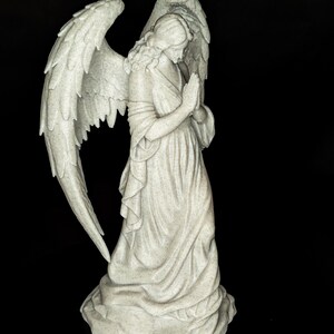 May include: A detailed statue of an angel with large, textured wings and a flowing gown. The angel is depicted with hands clasped in prayer, and the statue has a speckled, off-white finish. The background is solid black.