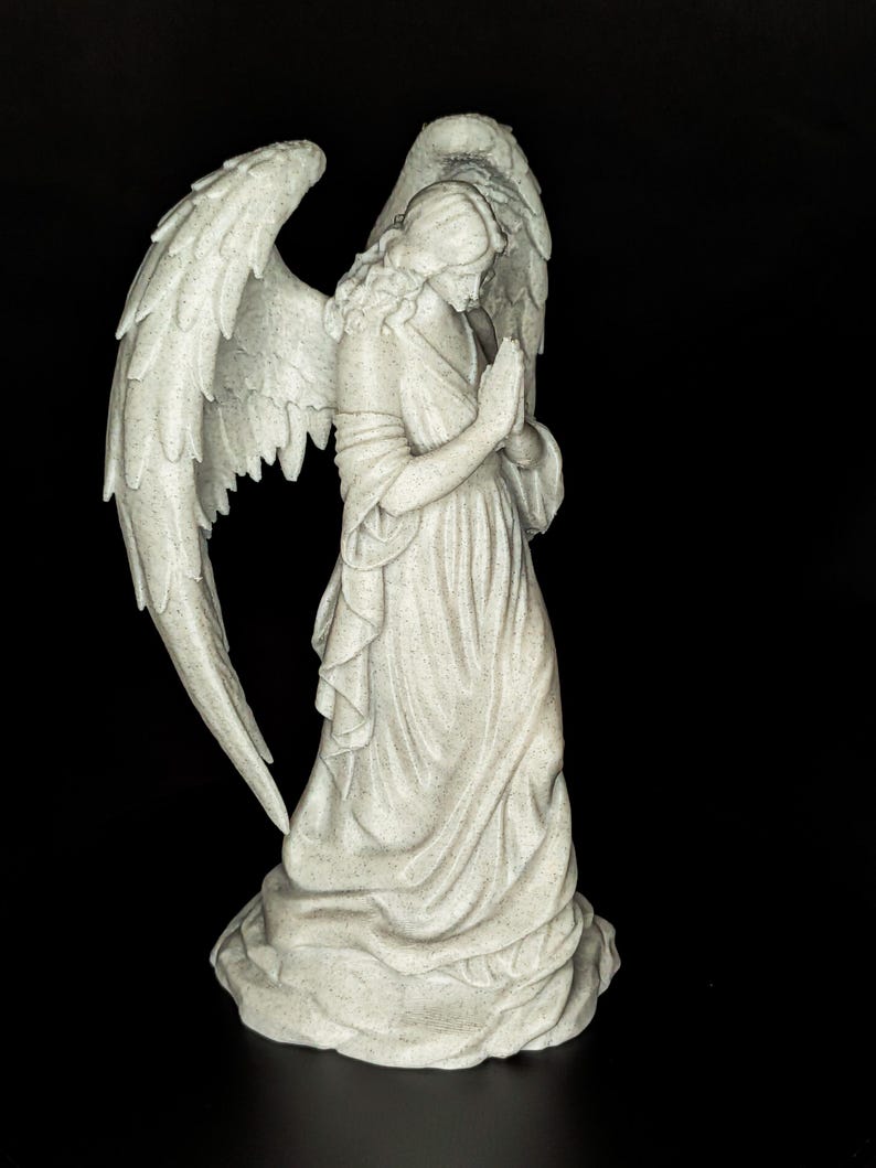 May include: A detailed statue of an angel with large, textured wings and a flowing gown. The angel is depicted with hands clasped in prayer, and the statue has a speckled, off-white finish. The background is solid black.