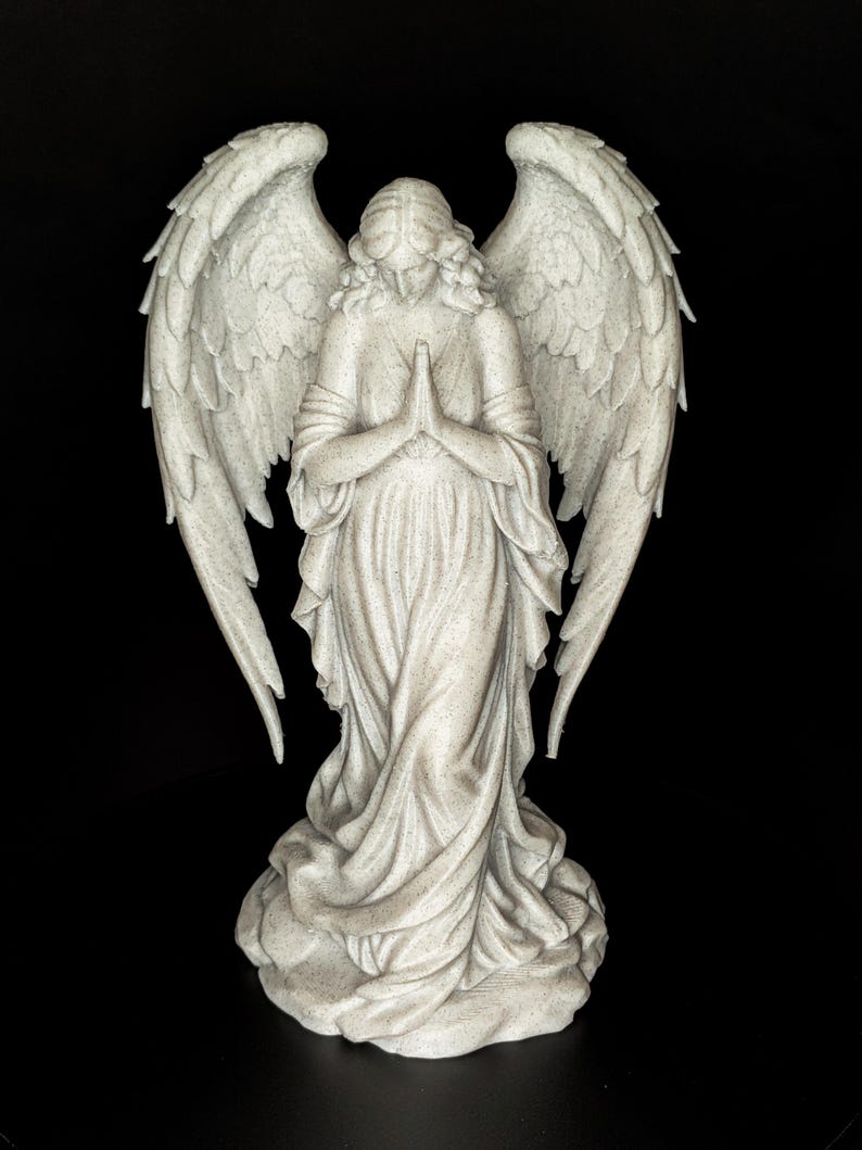 May include: A detailed statue of an angel with outstretched wings, hands clasped in prayer, and flowing robes. The sculpture is a light gray color and stands on a textured base. The background is solid black.