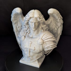 May include: A detailed bust of an angel with large, textured wings. The sculpture is light gray and features intricate details in the hair, armor, and a cross on the chest. The angel is set against a dark background.
