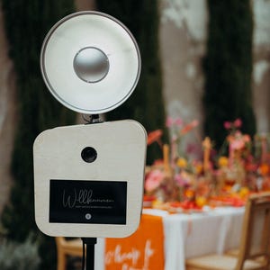 Peut inclure: Un photomaton avec une lumière circulaire brillante et un cadre en bois. Le photomaton a un écran noir avec le mot "Willkommen" en écriture blanche. Le photomaton est installé à l'extérieur avec une table et des chaises en arrière-plan.