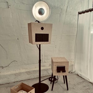 Peut inclure: Un photomaton en bois avec une lumière circulaire brillante, une petite boîte en bois et une caisse en bois. Le photomaton a un écran noir et est monté sur un support noir. La petite boîte a des pieds noirs.