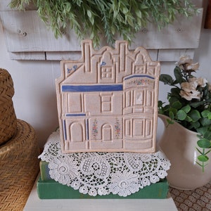 May include: A ceramic house-shaped napkin holder with a beige base, blue trim, and floral accents. The holder is sitting on a stack of books with a lace doily on top.