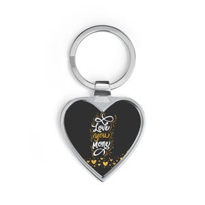 May include: Heart-shaped keychain with a black center and the words "Love You More" in white script. The keychain has a silver-colored metal ring and a border. Gold heart accents are at the bottom.