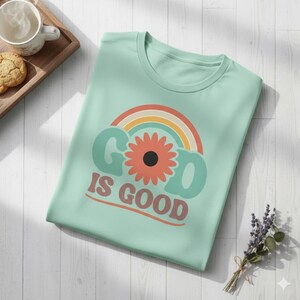 Puede incluir: Una camiseta verde claro con las palabras "GOD IS GOOD" en una fuente retro. El diseño incluye un arcoíris y una flor con un centro negro. La camiseta está doblada cuidadosamente sobre una superficie de madera blanca.