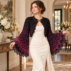 Peut inclure: Un boléro en crochet à manches longues présentant un dégradé de couleurs allant du noir aux épaules au violet profond et magenta sur les poignets évasés. Le vêtement possède un motif texturé en écailles et une attache métallique au col.