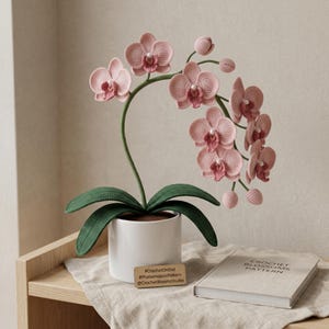 Op de afbeelding: Een gehaakte Phalaenopsis orchidee in een witte pot. De roze bloemen hebben gedetailleerde bloemblaadjes en een parelachtig centrum. De groene bladeren en stengel zorgen voor contrast. Een klein houten label en een boek zijn ook zichtbaar.