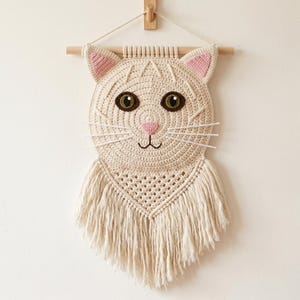 May include: A cream-colored, crocheted cat wall hanging. The cat's face features pink-lined ears, green eyes, and a pink nose. White yarn whiskers extend from the face. The cat's body has a macrame fringe. The wall hanging is suspended from a wooden dowel.