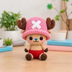 May include: A crochet amigurumi figure of a character with a tan face, black eyes, a blue nose, and pink cheeks. It wears a large pink hat with a white cross emblem and brown antlers, a tan torso, and red shorts with dark brown hands and feet.