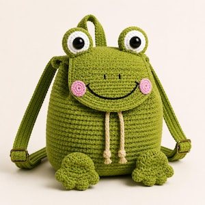 Puede incluir: Una mochila de ganchillo verde con forma de rana, con grandes ojos negros y blancos, mejillas circulares rosas y una sonrisa bordada. La bolsa cuenta con dos correas ajustables con hebillas de metal color bronce y dos cordones beige.