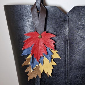 May include: A black textured tote bag featuring a decorative leather charm composed of multiple layered maple leaf cutouts in red, blue, yellow, and brown, attached to a dark brown leather strap with a small brass-colored metal rivet.