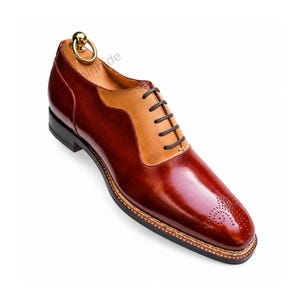 Pode incluir: Um sapato oxford de dois tons com corpo em couro vermelho escuro polido e painel de atacadores em couro bege. O sapato tem atacadores castanhos escuros, um padrão perfurado decorativo na biqueira, sola preta e uma forma de metal dourado.