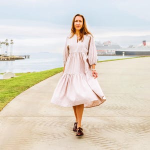 Può includere: Una persona che indossa un abito rosa chiaro con scollo a V e maniche lunghe cammina su un sentiero lastricato. L'abito ha una gonna a balze ed è abbinato a scarpe marrone scuro. Lo sfondo include acqua, palme e una grande nave.