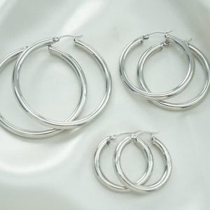 May include: Three pairs of silver hoop earrings are arranged on a white, silky background. The earrings are of different sizes, with a polished, reflective surface. The largest pair is at the top, followed by a medium and a small pair of hoops.