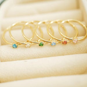 May include: Six gold-colored rings with small, colorful gemstones. The gemstones are blue, white, green, blue, orange, and pink. The rings are displayed in a beige jewelry box.