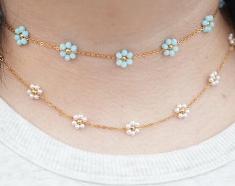 May include: Two gold-toned chain necklaces worn as chokers. The top necklace features light blue flower-shaped beads with a gold center. The bottom necklace features white flower-shaped beads with a gold center, creating a layered floral design.