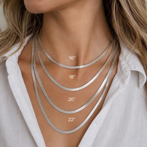 May include: Four silver-toned herringbone chain necklaces layered together. The lengths are labelled as 16 inches, 18 inches, 20 inches, and 22 inches, indicating the specific size of each individual chain in this set of flat, flexible metal jewellery.