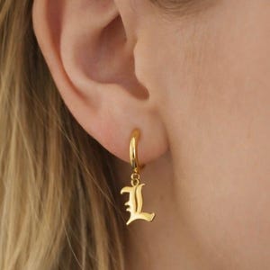 May include: Gold-colored hoop earring with a dangling letter "L" in an old English font. The earring is worn on a person's ear, with blonde hair visible in the background. The jewelry is a simple yet elegant accessory.