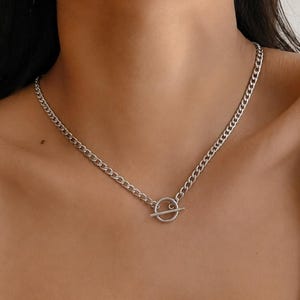 K&ouml;nnte beinhalten: Eine silberfarbene Kettenhalskette mit einem Knebelverschluss. Die Halskette besteht aus einer Reihe miteinander verbundener ovaler Glieder, die eine durchgehende Kette bilden, mit einem kreisf&ouml;rmigen Ring und einem horizontalen Stab vorne.