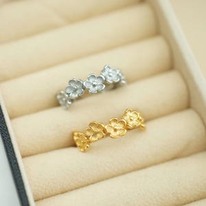 May include: Two rings featuring a series of small, five-petaled flowers connected in a band. One ring is finished in a bright silver tone, while the other ring is finished in a warm gold tone. Both rings have a textured, matte surface finish.