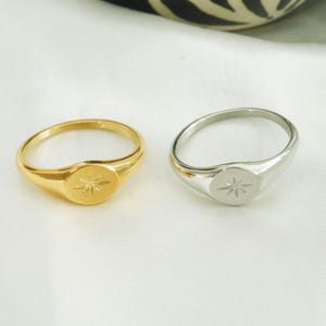 May include: Two signet rings, one gold-colored and one silver-colored, each with a starburst design on the oval face. The rings are displayed on a white, textured surface.