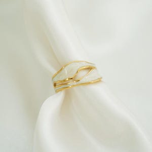 May include: A gold ring featuring a wavy, organic design with smooth white enamel inlay. The band consists of multiple interconnected, flowing gold lines that create an open, layered appearance, showcasing a polished metallic finish.
