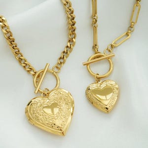 May include: Two gold necklaces with heart-shaped lockets. One has a chunky chain, the other a delicate chain. Both feature a toggle clasp. The necklaces are displayed on a white, silky background.