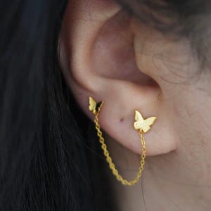 May include: Gold butterfly earrings. One butterfly is attached to the earlobe, and the other is connected by a delicate chain. The earrings are a bright gold color and are worn on the ear.
