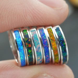 May include: A collection of silver rings with colorful opal inlays. The rings display a spectrum of colors, including blue, green, and orange. The rings are held between two fingers, showcasing the variety of designs.