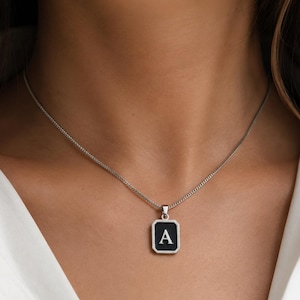 May include: A silver-toned necklace featuring a rectangular pendant with a black enamel inlay and a prominent silver letter A centered on the face. The pendant hangs from a delicate silver-toned chain that rests against the collarbone area.
