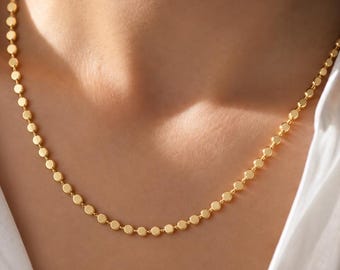 Könnte beinhalten: Eine goldfarbene Kettenhalskette, die aus einer Reihe kleiner, gleichmäßiger, flacher runder Scheiben besteht, die miteinander verbunden sind. Die Kette liegt auf einem weißen Hemd mit Kragen und zeigt ein gleichmäßiges metallisches Finish.