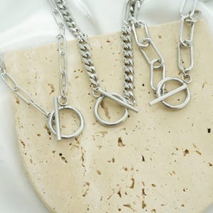 K&ouml;nnte beinhalten: Drei silberfarbene Metallhalsketten mit Knebelverschl&uuml;ssen. Jede Halskette besteht aus einem kreisf&ouml;rmigen Ring und einem T-Steg-Verschluss. Die Ketten variieren im Stil, darunter l&auml;ngliche ovale Glieder und engere Panzerkettenglieder.