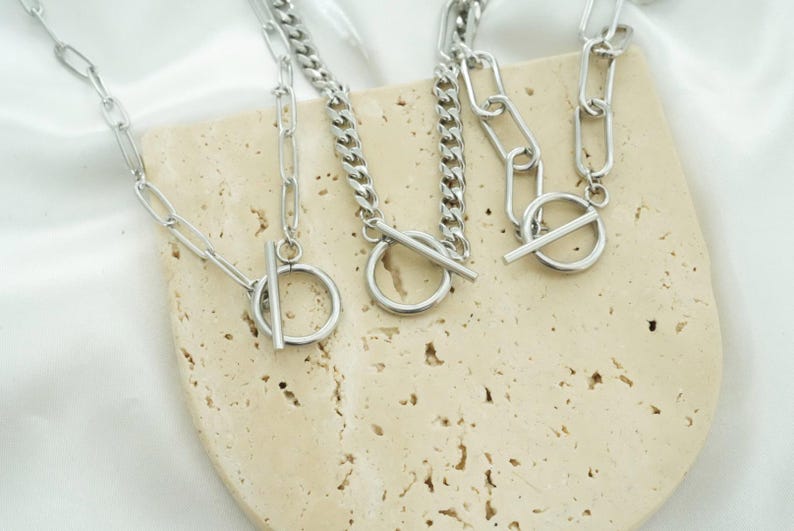 K&ouml;nnte beinhalten: Drei silberfarbene Metallhalsketten mit Knebelverschl&uuml;ssen. Jede Halskette besteht aus einem kreisf&ouml;rmigen Ring und einem T-Steg-Verschluss. Die Ketten variieren im Stil, darunter l&auml;ngliche ovale Glieder und engere Panzerkettenglieder.