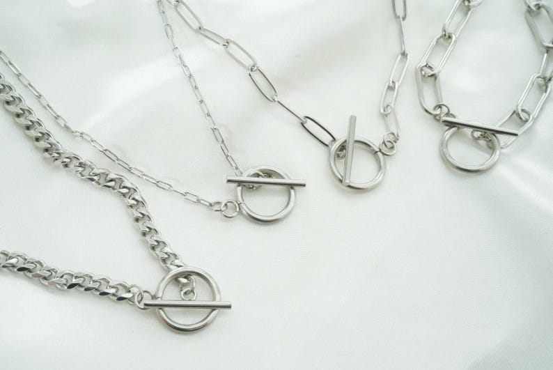 May include: Four silver necklaces with toggle clasps are displayed on a white surface. The necklaces vary in chain style, including a curb chain and a chain with elongated links. The clasps are circular with a straight bar.