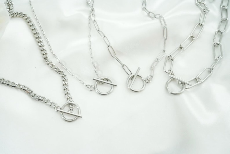 May include: Three silver-toned metal necklaces featuring distinct chain link styles. Each necklace is finished with a circular toggle clasp closure. The chains vary in thickness and link shape, including a curb chain and elongated oval links.