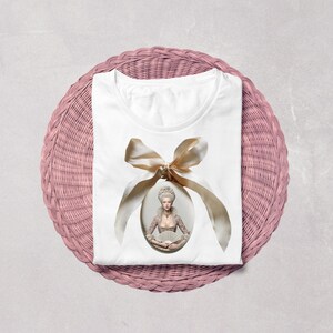 May include: A white t-shirt featuring a central oval graphic of a person in 18th-century attire with a powdered wig, holding a fan. The portrait is framed by a large, light beige satin ribbon tied in a bow at the top, centred on the chest.