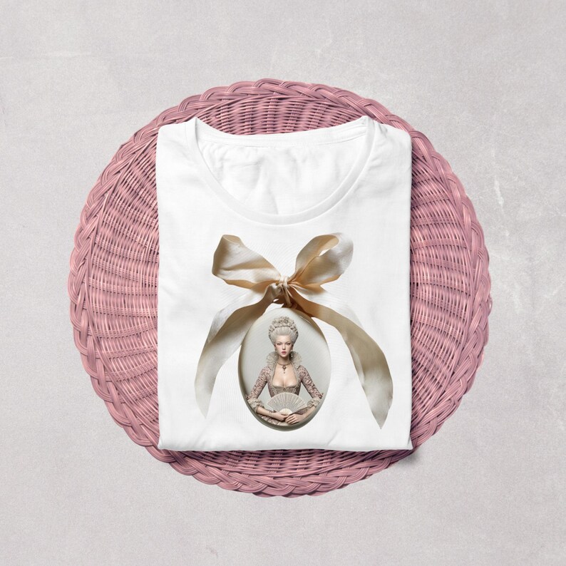 May include: A white t-shirt featuring a central oval graphic of a person in 18th-century attire with a powdered wig, holding a fan. The portrait is framed by a large, light beige satin ribbon tied in a bow at the top, centred on the chest.