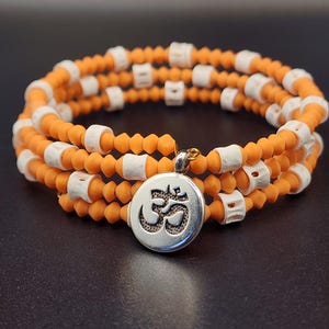 Puede incluir: Una pulsera envolvente con cuentas naranjas y detalles rectangulares blancos. Un dije plateado con el símbolo Om cuelga. La pulsera se muestra sobre una superficie oscura.