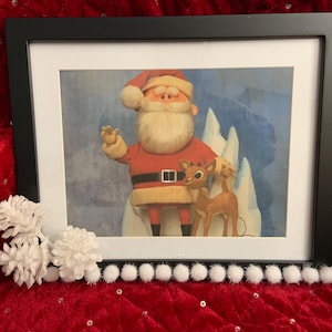 Könnte beinhalten: Gerahmtes Weihnachtsbild mit dem Weihnachtsmann und einem Rentier vor einer verschneiten Kulisse. Der schwarze Rahmen hat weiße Pompons und weiße Blumendekorationen. Das Bild ist auf einer roten, paillettenbesetzten Oberfläche platziert.