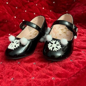 Peut inclure: Paire de chaussures Mary Jane noires en cuir verni, ornées de flocons de neige et de pompons blancs. Les chaussures ont une fermeture à boucle et sont posées sur un fond rouge.