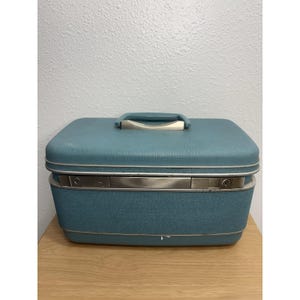 May include: A rectangular, light blue hardshell train case with a textured exterior finish. It features a silver-toned metal latch mechanism across the front and a matching light blue handle with silver accents on the top lid.