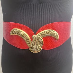 Puede incluir: Un cinturón ancho de color rojo con una textura similar a la piel de serpiente y una hebilla grande de metal dorado. La hebilla presenta un diseño curvo con patrón de espiga que se entrelaza en el centro, creando un punto focal llamativo.