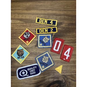 May include: A collection of embroidered patches, including "DEN 4", "DEN 2", "WOLF", "BOBCAT", "BEAR", and "QUALITY UNIT 1999". The patches feature various colors and designs, with images of animals and numbers.