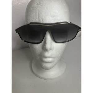 May include: Black sunglasses featuring a thick, angular frame with a prominent brow bar. The lenses have a dark gradient tint, and the bridge area is accented with a thin, horizontal gold-toned metal strip that extends across the top of the frame.