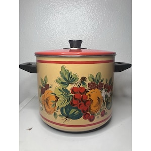 May include: A large cylindrical stockpot with a cream-colored body and a red lid. The pot features a decorative wrap-around illustration of various fruits and green leaves in shades of red, orange, and dark green, with two black side handles.