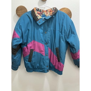 May include: A vintage teal and pink jacket with a geometric design. The jacket features a button-up front, two pockets, and a patterned collar. The jacket is hanging on a white hanger against a white wall.