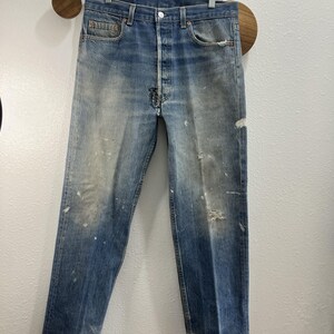 May include: A pair of light blue denim jeans featuring a button fly closure, five pockets, and a distressed finish with visible white paint splatters, fading, and a small frayed tear on the right knee area, showcasing a worn-in aesthetic.